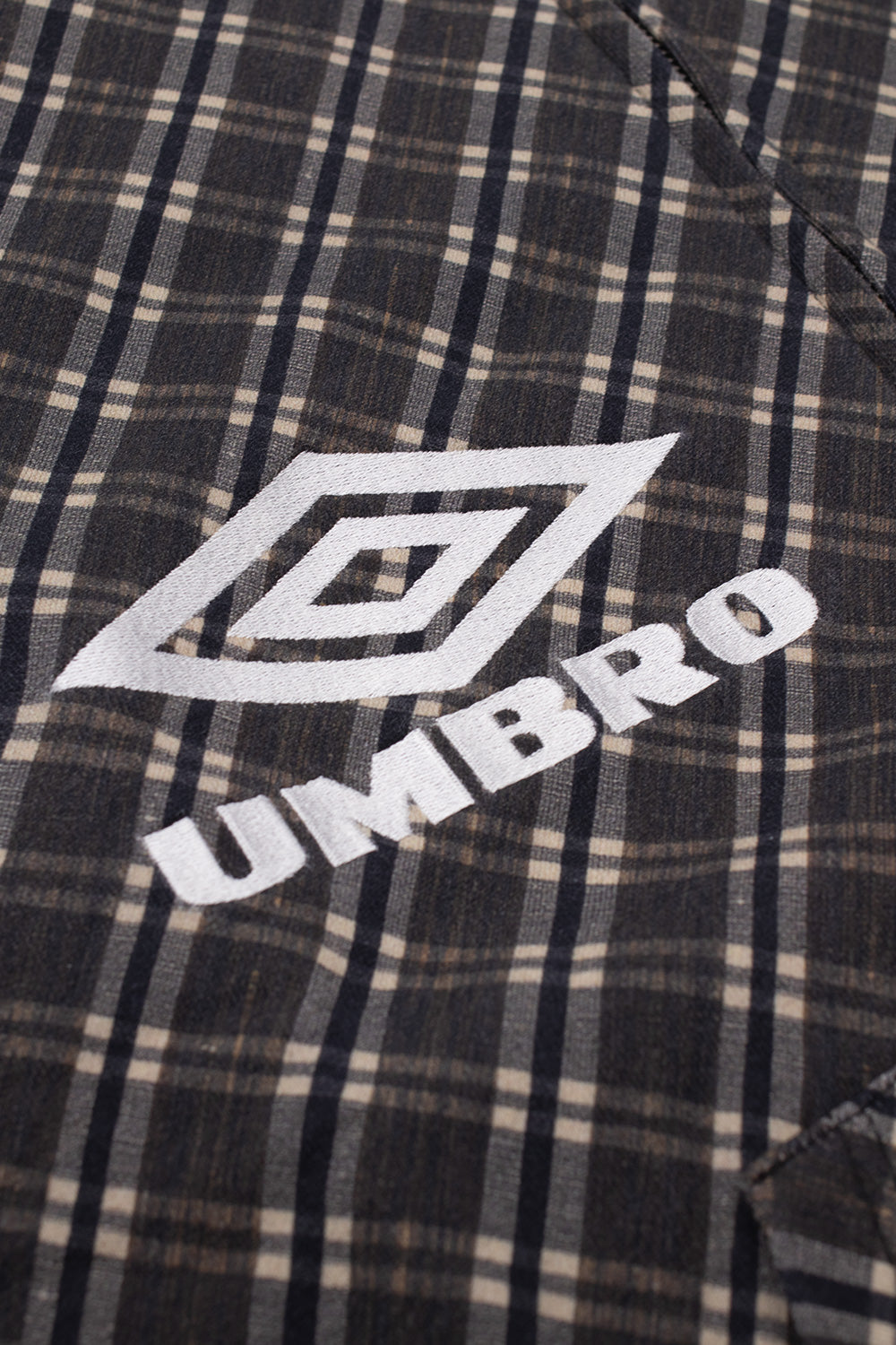 Close-up shot of the chest embroidery on the brown Umbro Plaid Masked Hoodie, highlighting the white satin-stitched Umbro double diamond logo and serif brand lettering on the woven brown and beige check textile