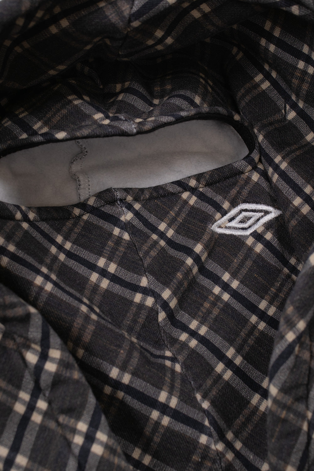 Detailed interior view of the hood on the Umbro Plaid Masked Hoodie, displaying the integrated brown plaid face mask panel with a bound eye slit opening and a small white embroidered Umbro double diamond logo on the cheek