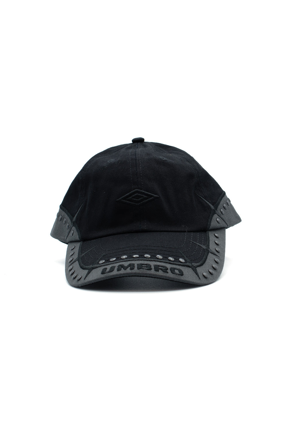 Front studio portrait of the Umbro Spikes Stud Cap in washed black cotton twill, featuring a low-profile 6-panel crown, tonal black satin-stitched Umbro double diamond front embroidery, and a dark charcoal contrast visor edge bordered with matte gunmetal domed metal studs, sharp spiked star embroidery, and a bold arched Umbro wordmark across the curved brim