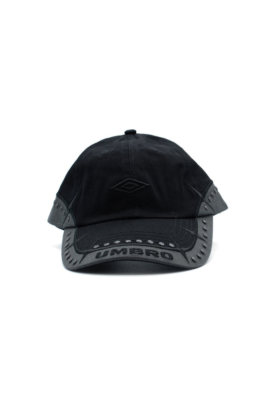 Front studio portrait of the Umbro Spikes Stud Cap in washed black cotton twill, featuring a low-profile 6-panel crown, tonal black satin-stitched Umbro double diamond front embroidery, and a dark charcoal contrast visor edge bordered with matte gunmetal domed metal studs, sharp spiked star embroidery, and a bold arched Umbro wordmark across the curved brim