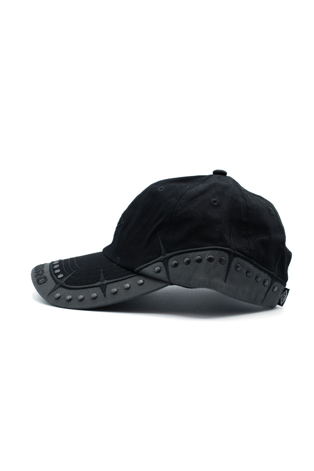 Left side profile shot of the Umbro Spikes Stud Cap, highlighting the dark grey scalloped canvas overlay wrapping from the visor along the entire bottom edge of the crown, studded with spaced metal dome hardware, accented by pointed black spike threadwork and an embroidered mini logo tag