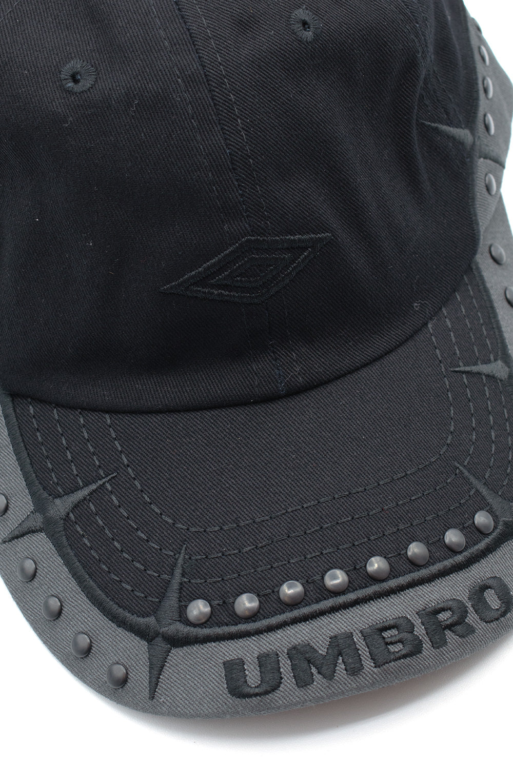 Macro close-up view of the visor and lower crown seam on the Umbro Spikes Stud Cap, displaying the thick black satin-stitched Umbro double diamond logo, curved row of matte gunmetal studs, and dense black dimensional embroidery forming barbed spikes and block brand lettering