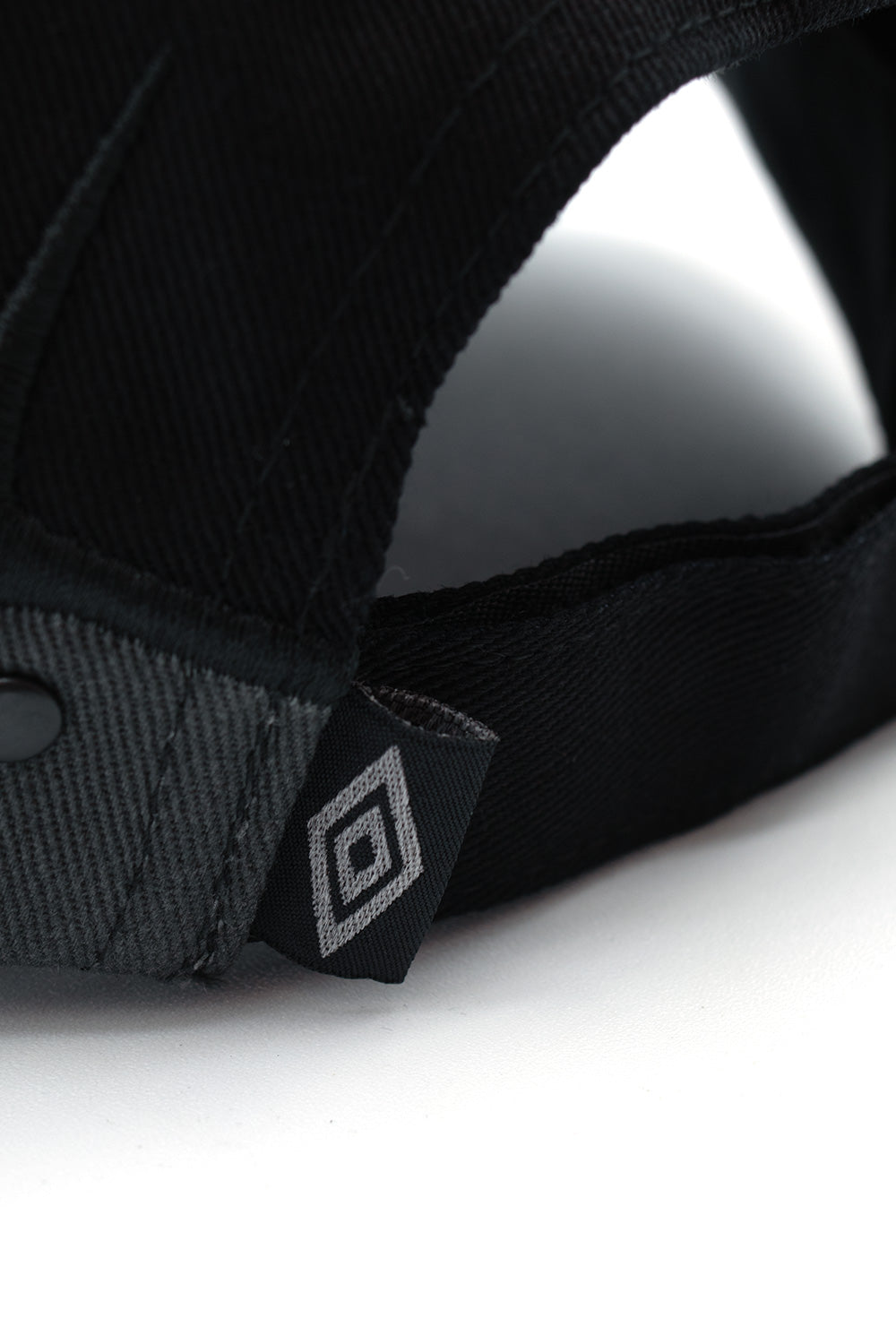 Detailed close-up shot of the rear closure strap on the Umbro Spikes Stud Cap, highlighting the woven black flag label featuring the silver-grey Umbro double diamond logo sewn securely alongside the edge-stitched cotton Velcro band