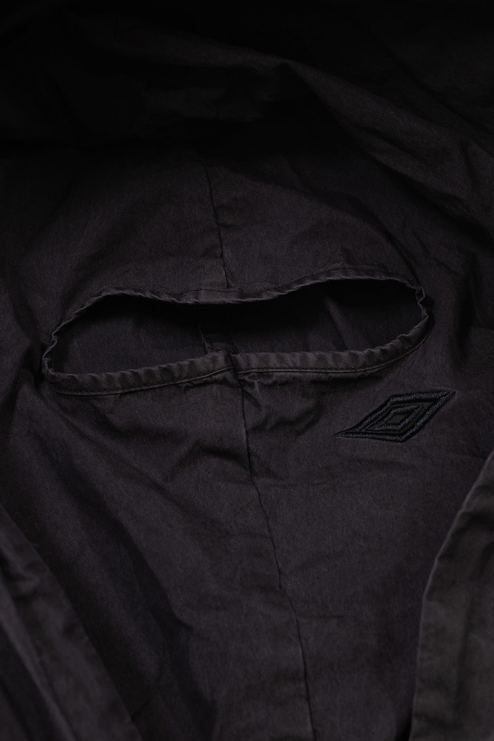 Detailed studio close-up of the hood on the Umbro Sunday Masked Jacket, highlighting the integrated washed black balaclava face mask panel with a bound eye slit opening and a subtle tonal embroidered Umbro double diamond logo on the cheek