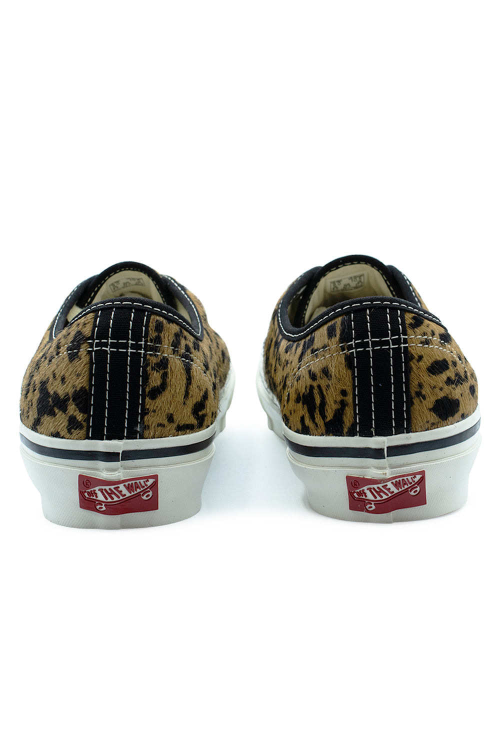 Rear view of the heels of the Vans LX Authentic 44 Cow Marshmallow sneakers, showing the textured cow-print pony hair heel counters with black canvas heel strip overlays, contrast white double stitching, and red vintage Off The Wall skateboard license plates on the white foxing tape.