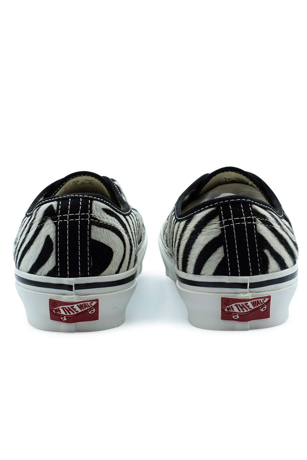 Rear view of the heels of the Vans LX Authentic 44 Zebra Marshmallow sneakers, displaying the textured zebra-print pony hair heel counters with black canvas heel strip overlays, contrast white double stitching, and red vintage Off The Wall skateboard license plates on the white foxing tape.