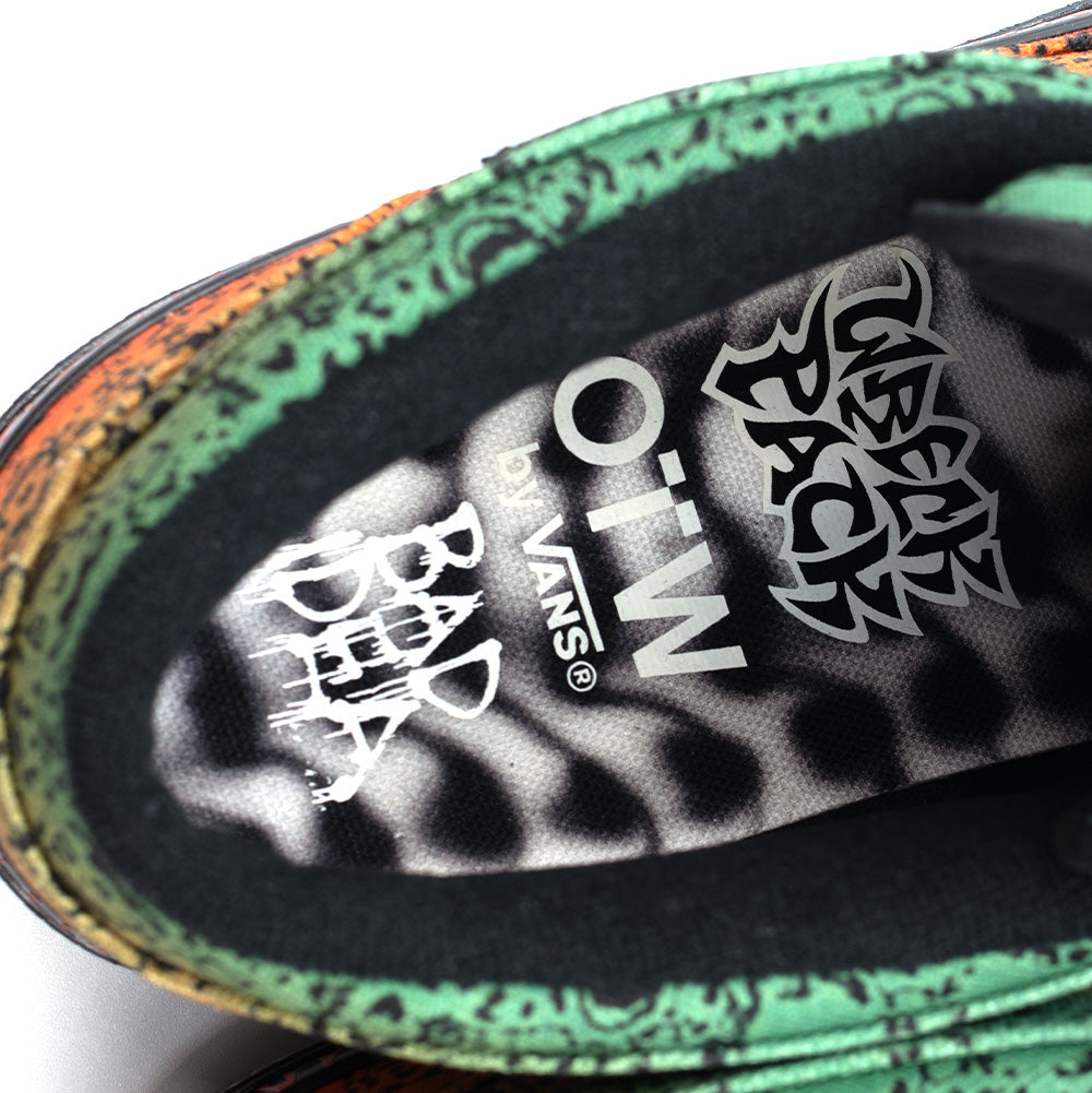 Extreme macro close-up looking down into the padded collar of the Vans OTW x Chito Half Cab 33 sneaker, displaying the cushioned insole printed with an airbrushed monochrome ribcage graphic, centered white "OTW by VANS" branding, dripping graffiti-style "BAD DEA" artwork, and silver metallic tribal script, framed by the green-and-orange skull-printed canvas collar rim.