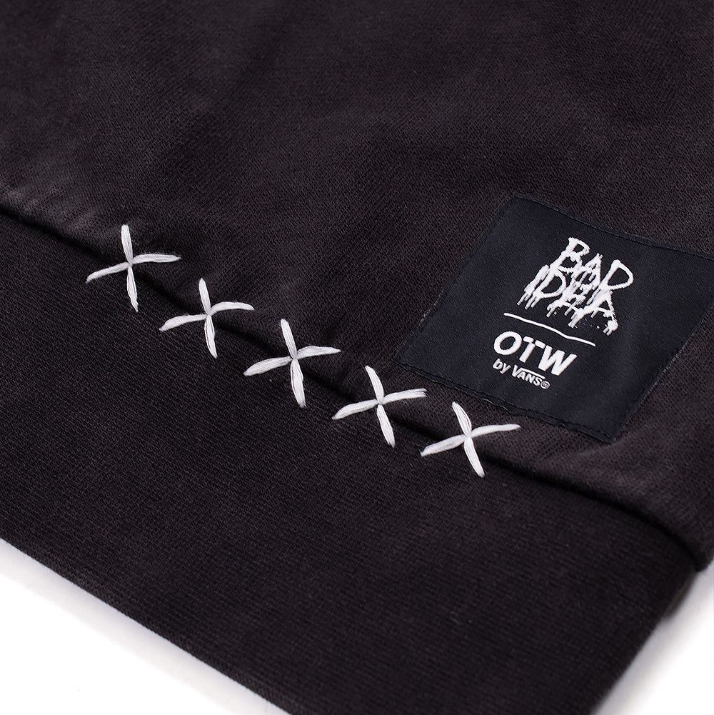 Macro close-up shot of the lower left hemline on the Vans OTW x Chito Sweatshirt, featuring a row of five thick white embroidered 'XXXXX' cross-stitches stitched beside a rectangular black woven label displaying Chito's "BAD IDEA" dripping graphics above the "OTW by VANS" logo.