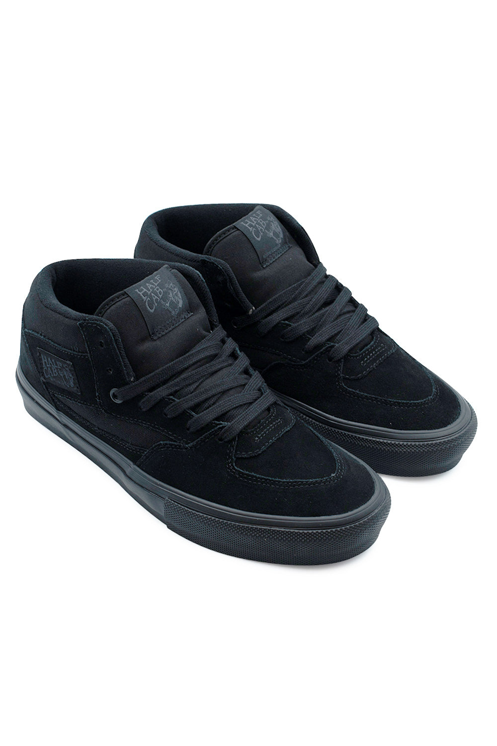 Elevated three-quarter studio view of the all-black Vans Skate Half Cab sneakers, emphasizing the iconic 1992 mid-top silhouette, hidden Duracap rubber underlay zones, impact-absorbing PopCush footbeds, and tonal black stitching.