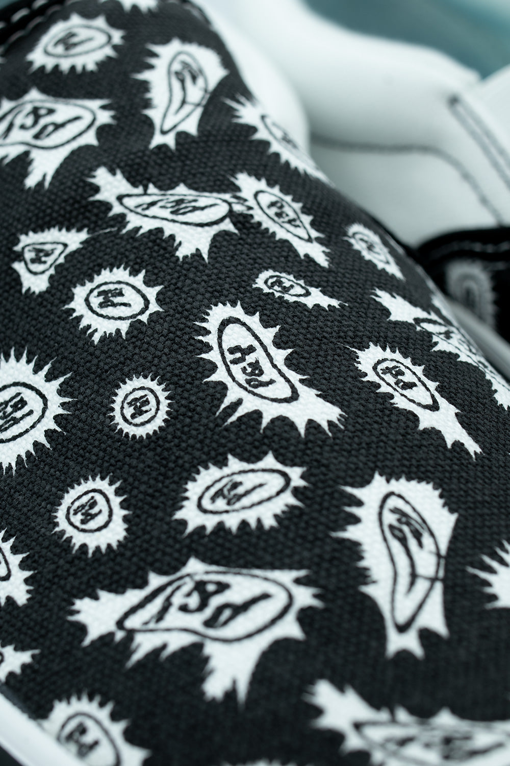 Detailed macro close-up of the woven black cotton canvas upper on the Vans X Punk And Yo Skate Slip-On, displaying the intricate white screen-printed spiked oval emblems containing stylized "P&Y" letters inspired by classic streetwear bandanas