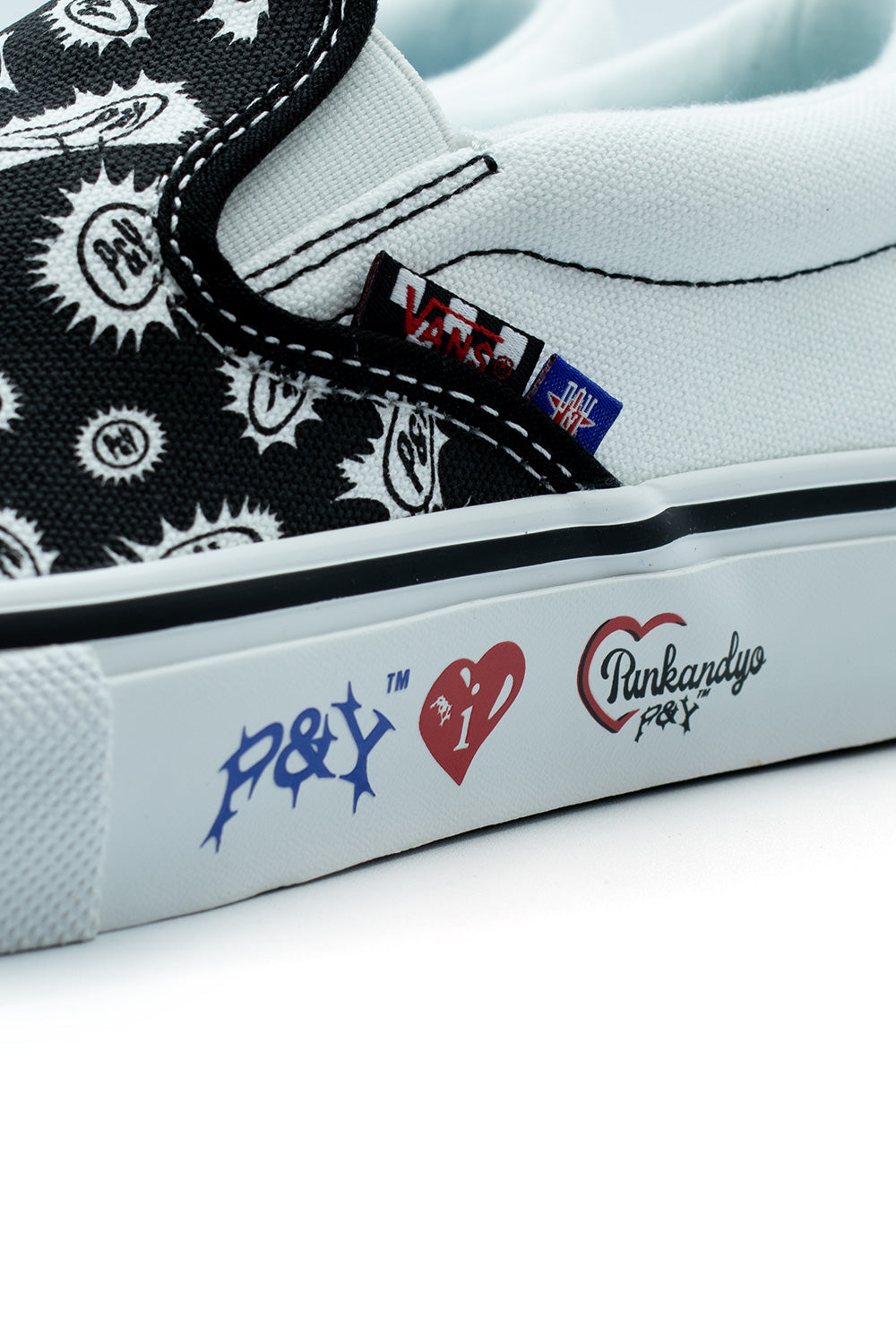 Macro studio view of the white vulcanized rubber midsole on the Vans X Punk And Yo Skate Slip-On, highlighting the crisp black foxing pinstripe and printed graphic logos including blue gothic "P&Y TM", a red heart with dagger motif, and retro "Punkandyo P&Y" script lettering