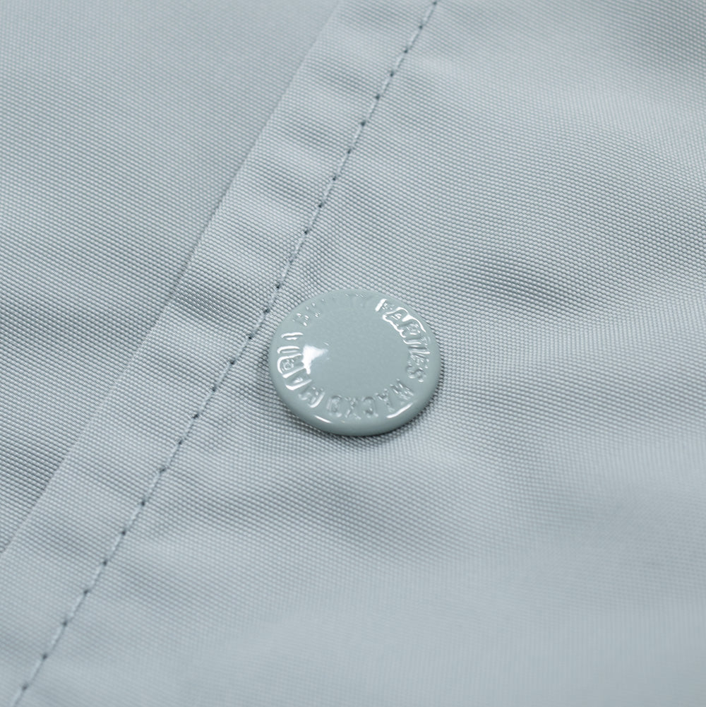 Close-up shot of the front placket closure on the Wacko Maria Coach Jacket Type 2, focusing on the light grey glossy metal snap fastener subtly debossed with circular "THE GUILTY PARTIES WACKO MARIA" branding set against the finely textured technical shell fabric.