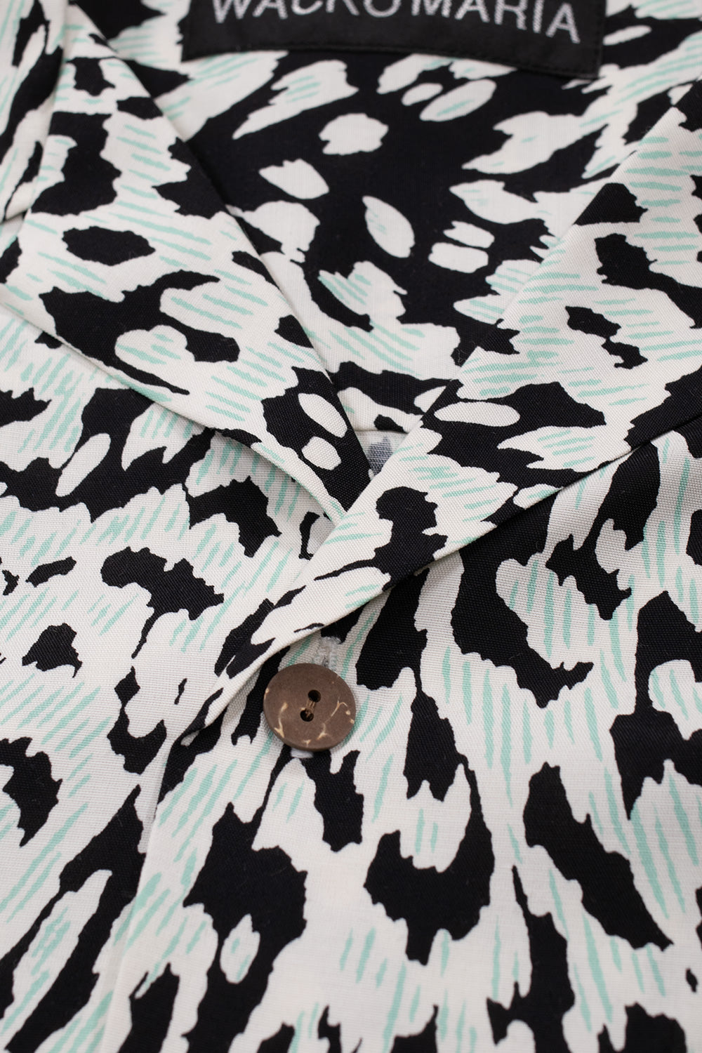 Close-up detail of the open camp collar and the top wooden button on the ivory Wacko Maria rayon Hawaiian shirt, showcasing the black and mint print texture.