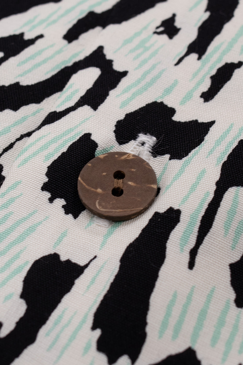 Macro view of a natural round wooden button stitched onto the patterned button placket of the ivory Wacko Maria rayon Hawaiian shirt.