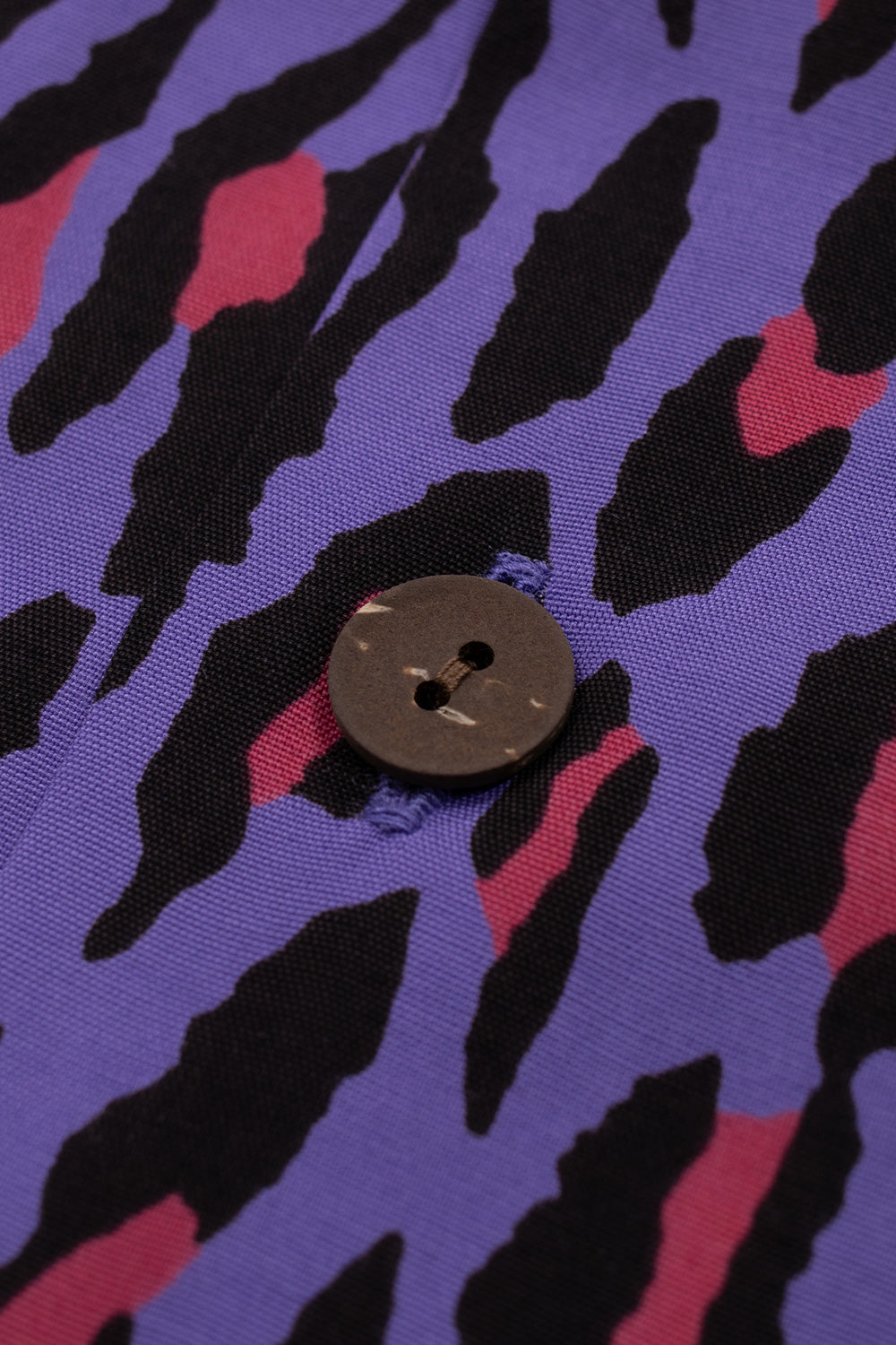 Macro view of a natural round wooden button stitched onto the button placket of the purple Wacko Maria rayon Hawaiian shirt.