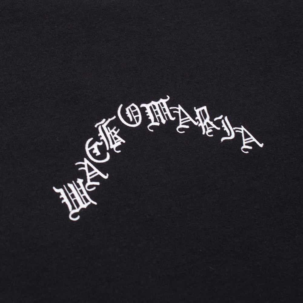 Macro perspective of the front left chest graphic on the black Wacko Maria T-Shirt, capturing the clean, opaque white ink finish of the curved Old English gothic "WACKO MARIA" typography printed onto vintage-washed heavyweight black cotton fabric.