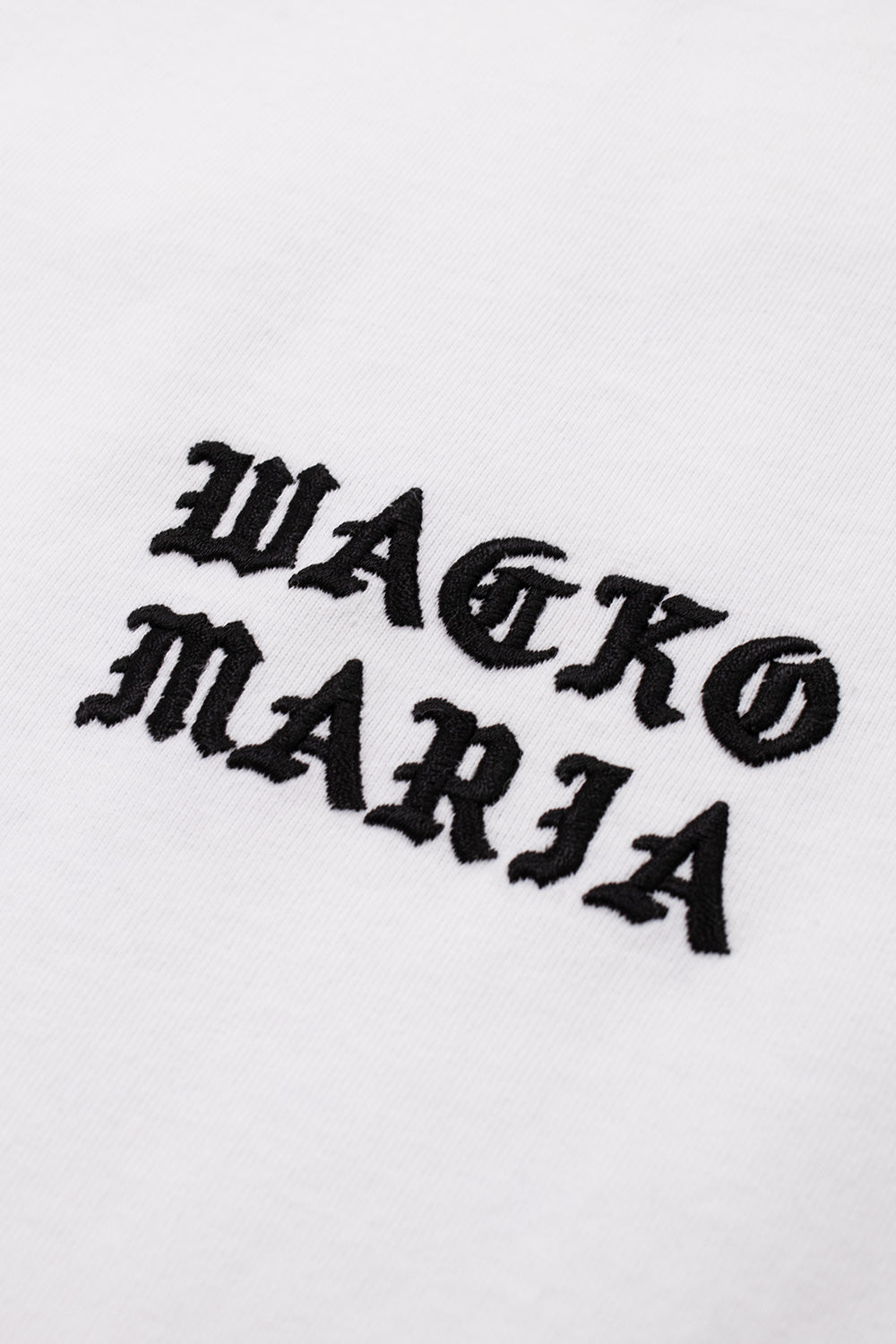 Detailed close-up of the black embroidered Wacko Maria Gothic font logo on the chest of the white t-shirt.
