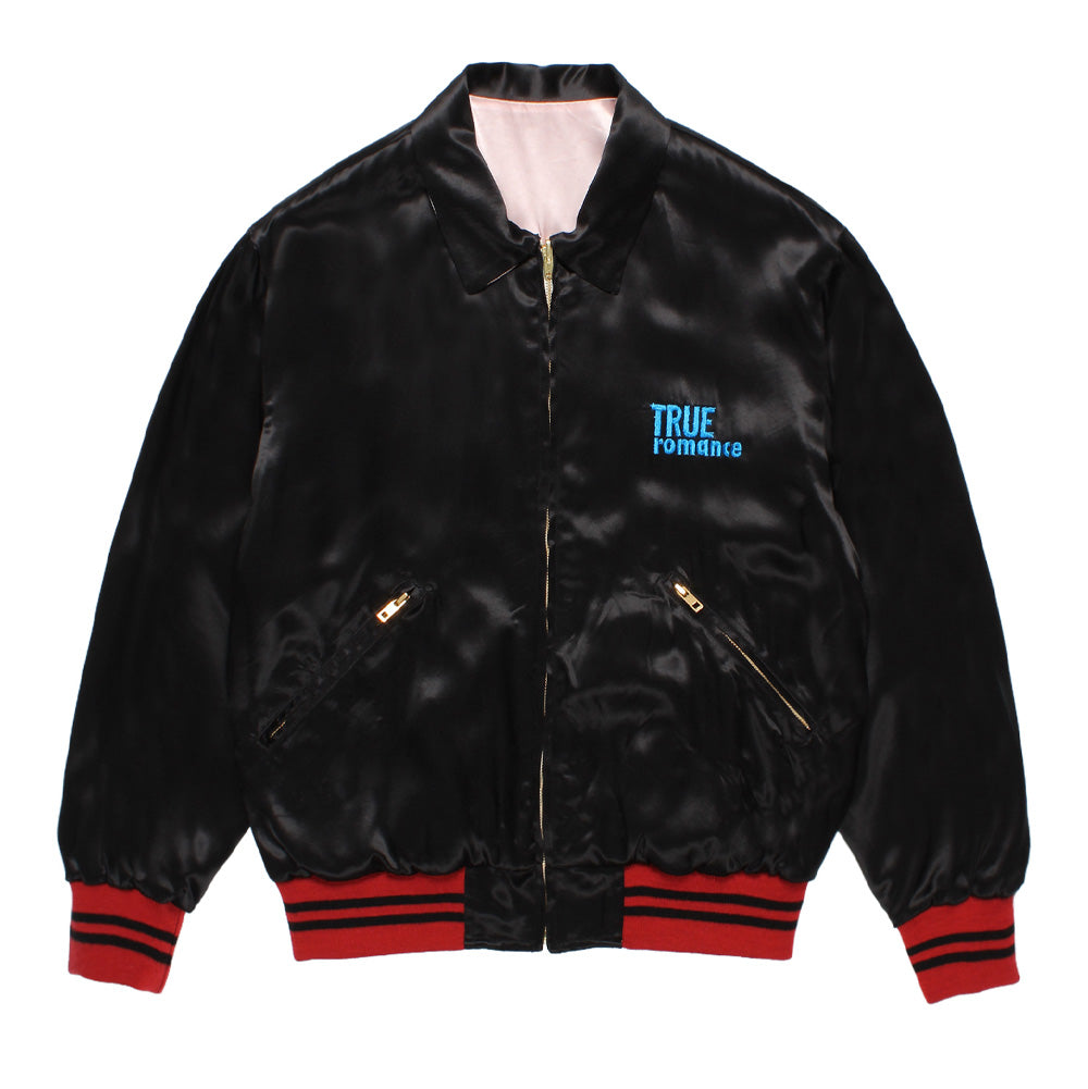 Front flat lay studio view of the black satin side of the Wacko Maria x True Romance Reversible Ska Jacket Type 2, showcasing a pointed turn-down collar, gold-tone metal center zipper closure, diagonal zippered welt pockets, two-tone red striped knit trims, and bright blue embroidered "TRUE ROMANCE" script on the left chest.