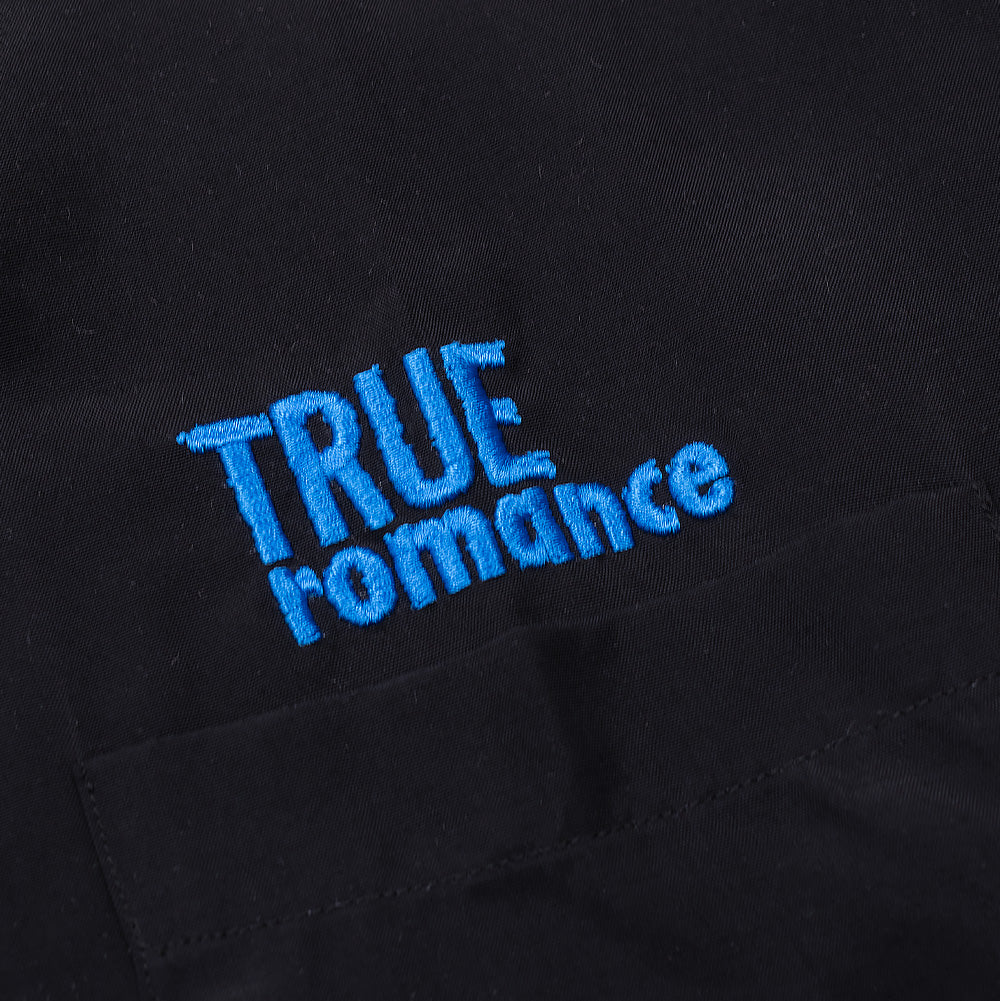 Detailed macro perspective of the chest branding on the Wacko Maria x True Romance Hawaiian Shirt, highlighting the rich turquoise-blue satin-stitch embroidery spelling out "TRUE ROMANCE" positioned cleanly above the front chest pocket on smooth black textile.