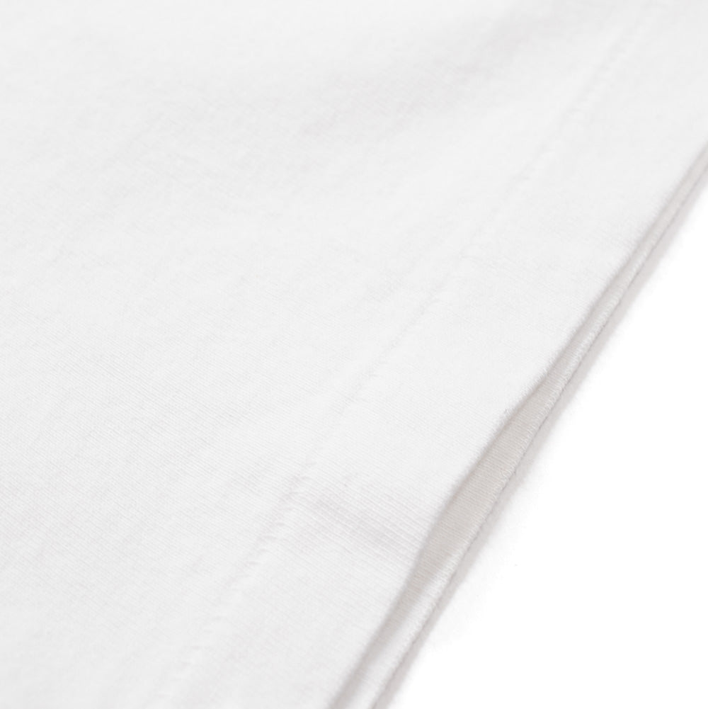 Extreme macro close-up of the hemline on the white Wacko Maria x True Romance Heavy Weight T-Shirt, focusing on the substantial knit texture, soft washed finish, and tonal double-stitched parallel edge seams of the premium heavyweight cotton fabric.